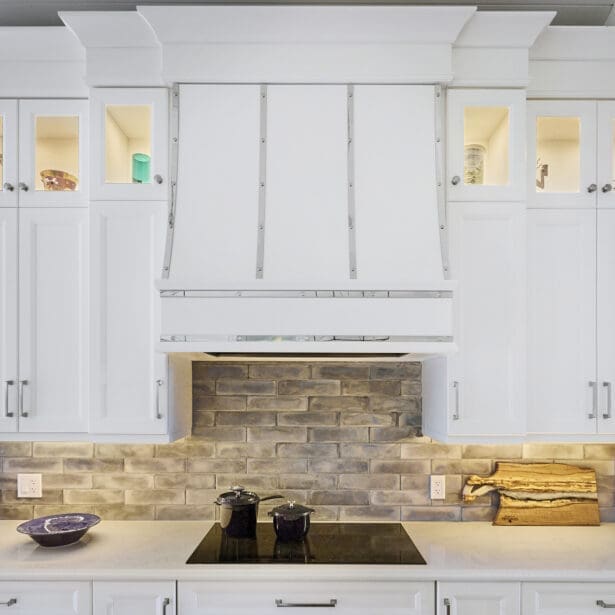 Interior view of a kitchen featuring white cabinetry, a range hood with chrome accents, an induction range, and a stylish brick backsplash, creating a modern and chic cooking space.