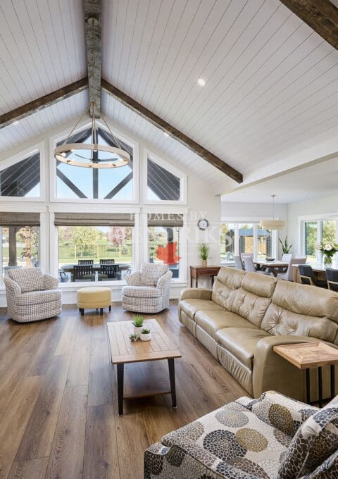 A grand great room featuring stunning timber accents that add warmth and rustic charm to the space. The high ceilings are framed by exposed wooden beams, while large windows allow natural light to flood the room