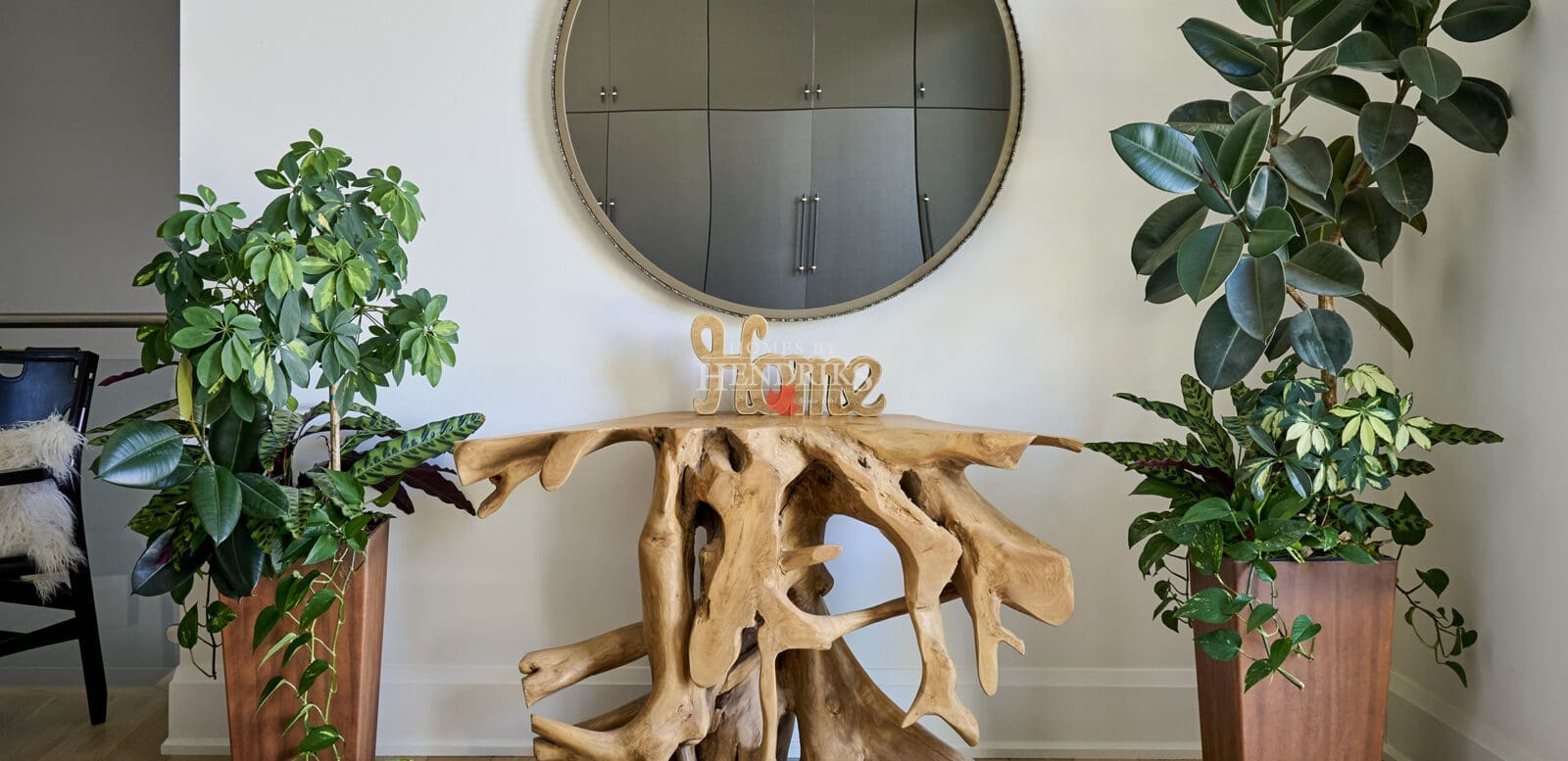 Modern entryway styled with a natural wood root console table, round mirror, and twin potted green plants in tall planters on either side