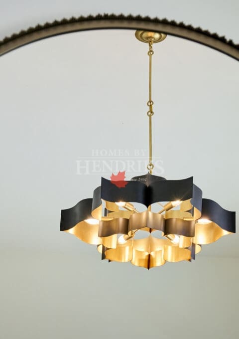 Modern gold and black chandelier viewed through a round mirror, featuring sculptural geometric metalwork and warm lighting in a minimalist white space
