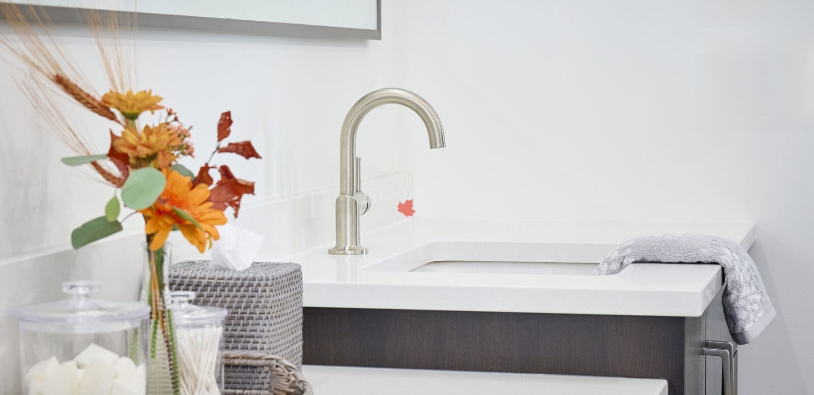 Close-up of a modern bathroom vanity faucet on a white quartz countertop with decorative tray, orange flowers, and textured accessories.