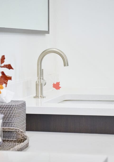 Close-up of a modern bathroom vanity faucet on a white quartz countertop with decorative tray, orange flowers, and textured accessories.