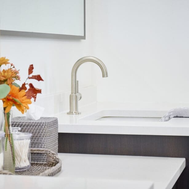 Close-up of a modern bathroom vanity faucet on a white quartz countertop with decorative tray, orange flowers, and textured accessories.
