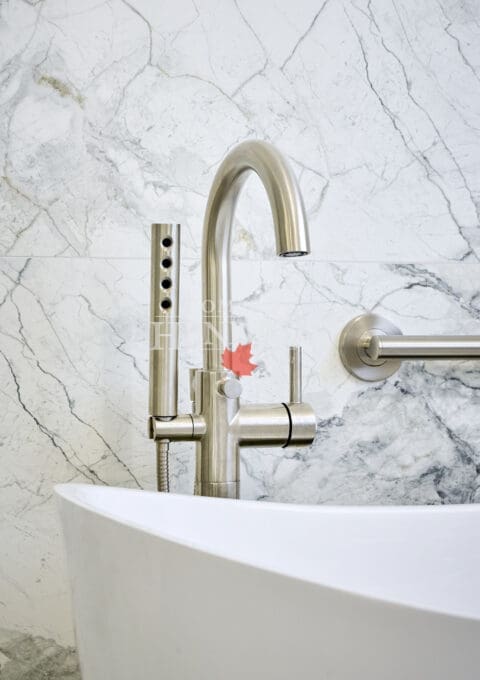 Close-up of brushed nickel bathtub faucet and handheld sprayer mounted on marble wall above a sculptural freestanding tub.