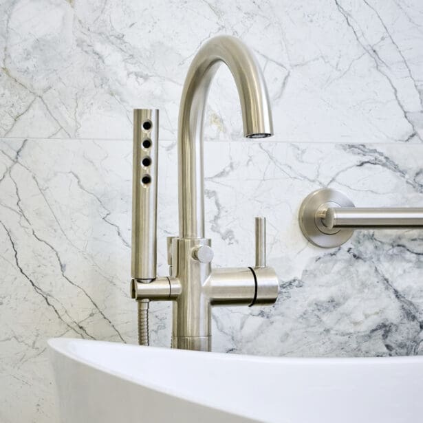 Close-up of brushed nickel bathtub faucet and handheld sprayer mounted on marble wall above a sculptural freestanding tub.