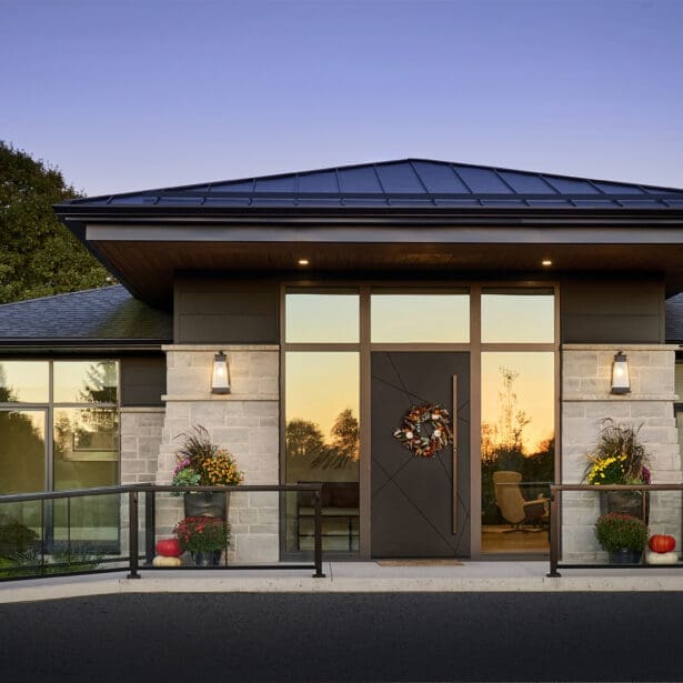 Modern single-story home with a low-slope metal roof, limestone accents, dark exterior trim, and large glass entryway at sunset.
