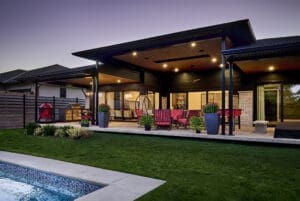 Covered outdoor living space with recessed lighting, red patio furniture, built-in grill station, and a view of the backyard pool at dusk.