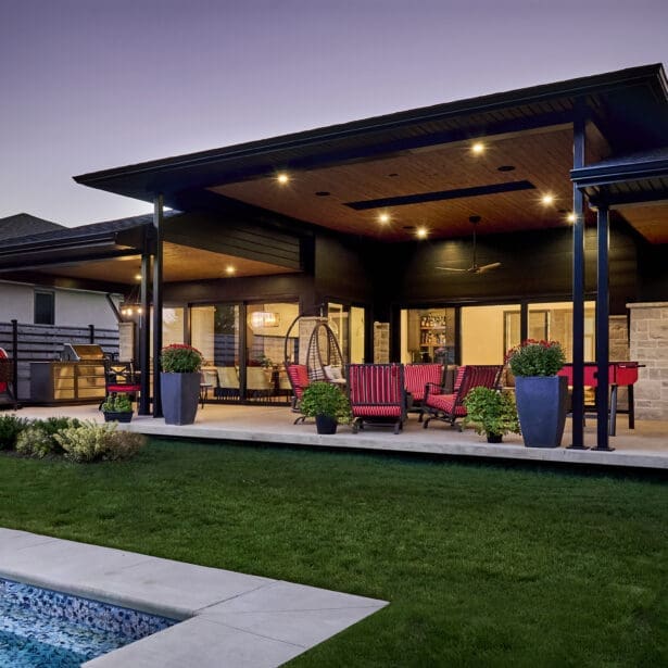 Covered outdoor living space with recessed lighting, red patio furniture, built-in grill station, and a view of the backyard pool at dusk.