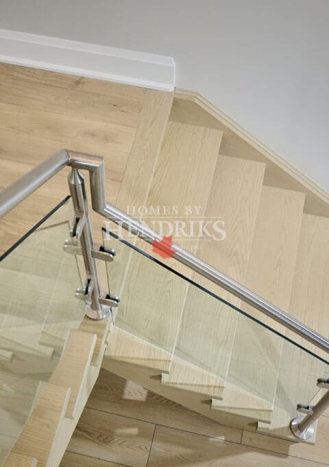 Staircase with light oak treads and glass railing framed by stainless steel hardware, viewed from above in a modern home interior.