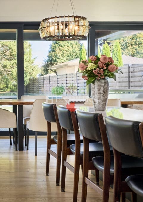 Bright dining area with large windows, wood flooring, modern chandelier, and a long dining table adorned with a floral centerpiece.