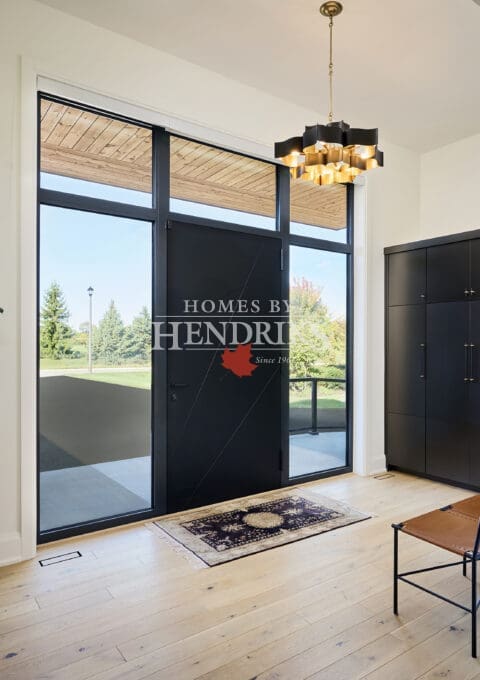 Spacious front entryway with tall black glass door, light hardwood floors, gold chandelier, and modern minimalist decor overlooking a green outdoor landscape