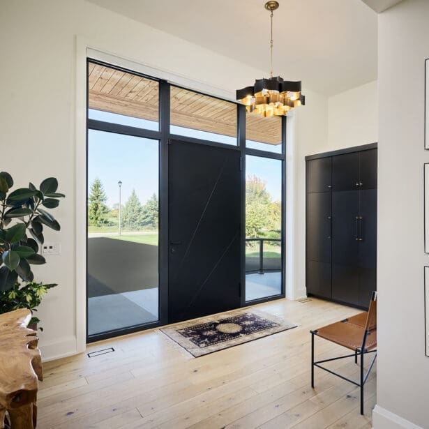 Spacious front entryway with tall black glass door, light hardwood floors, gold chandelier, and modern minimalist decor overlooking a green outdoor landscape