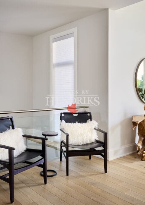Upper hallway seating area with two black armchairs, white fur pillows, and glass railing. Includes pendant light, sculptural wood console table, and circular mirror accent