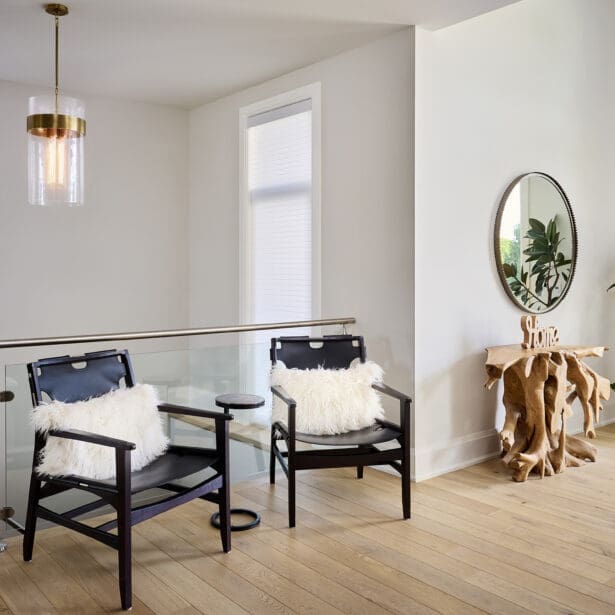 Upper hallway seating area with two black armchairs, white fur pillows, and glass railing. Includes pendant light, sculptural wood console table, and circular mirror accent