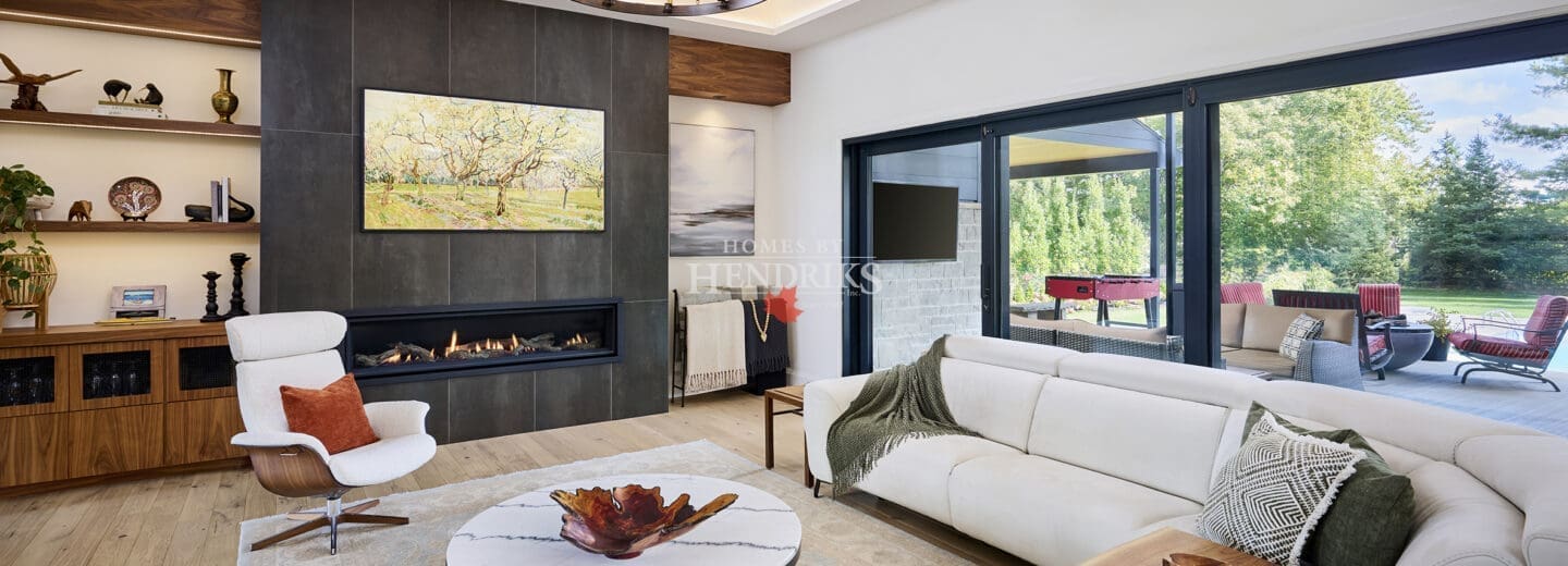 Bright contemporary living room featuring a sleek linear fireplace, modern chandelier, white sofas, and marble coffee table. Large windows open to a covered outdoor lounge