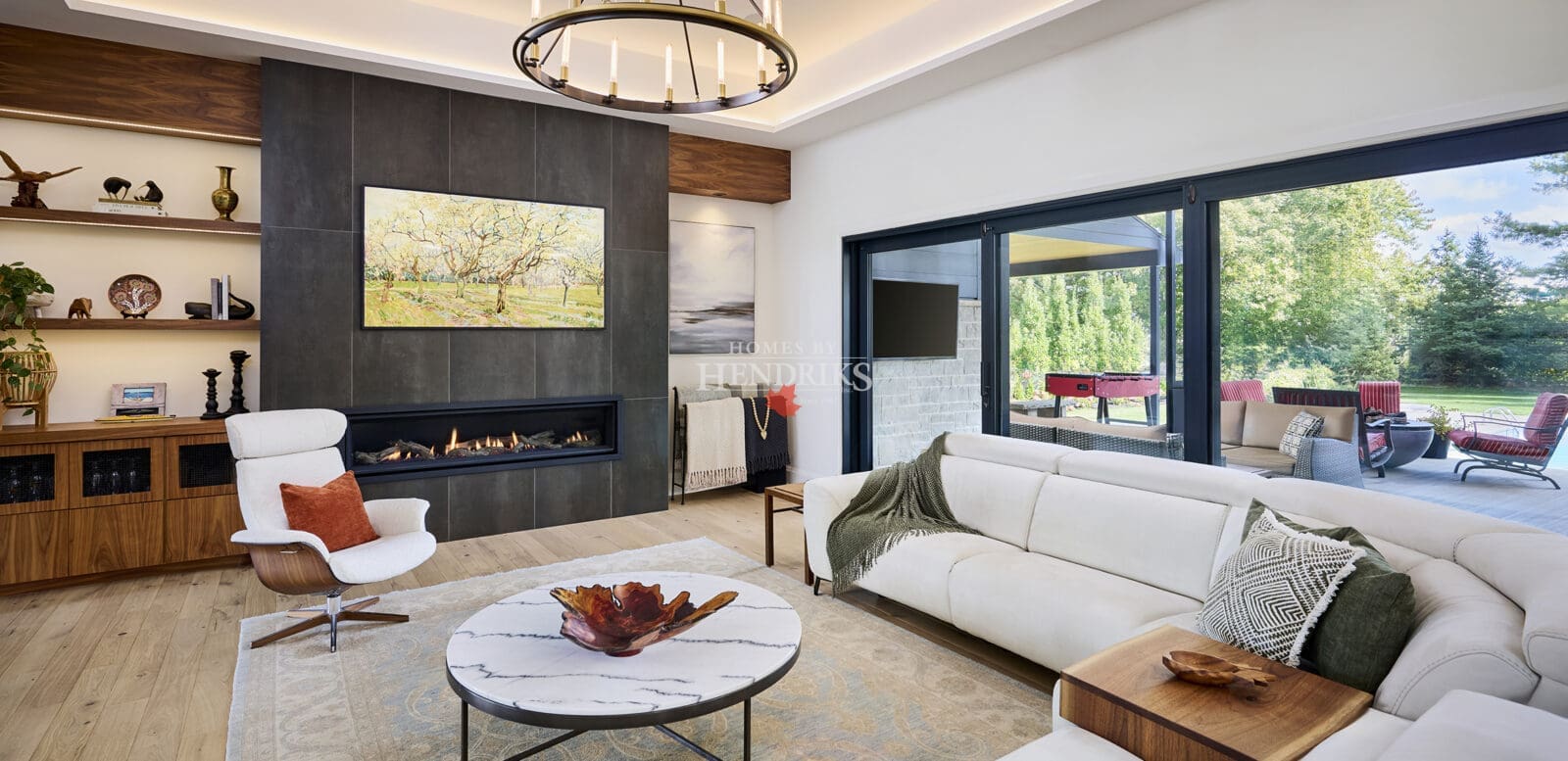 Bright contemporary living room featuring a sleek linear fireplace, modern chandelier, white sofas, and marble coffee table. Large windows open to a covered outdoor lounge