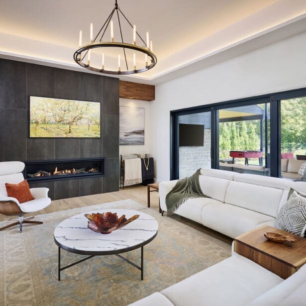 Bright contemporary living room featuring a sleek linear fireplace, modern chandelier, white sofas, and marble coffee table. Large windows open to a covered outdoor lounge