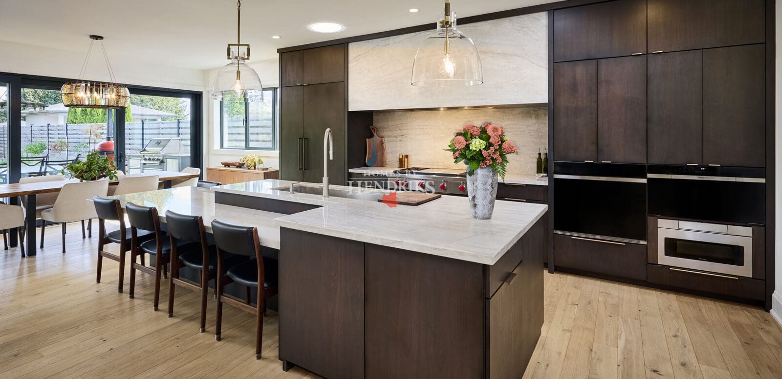 Modern open-concept kitchen and dining area featuring dark wood cabinetry, marble countertops, and large glass doors opening to an outdoor patio. Pendant lighting and floral accents add warmth and sophistication