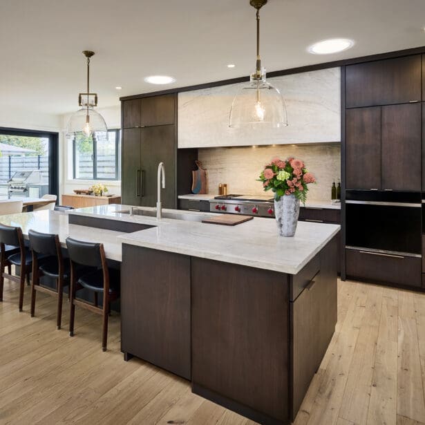 Modern open-concept kitchen and dining area featuring dark wood cabinetry, marble countertops, and large glass doors opening to an outdoor patio. Pendant lighting and floral accents add warmth and sophistication
