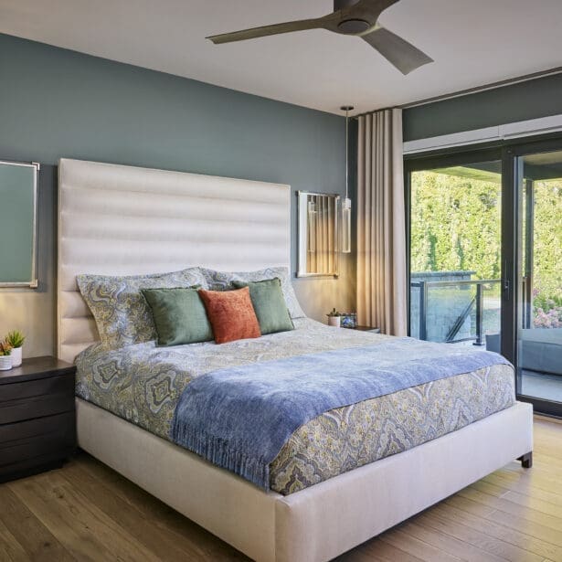 Contemporary bedroom with upholstered bed, patterned bedding, and accent pillows in green and terracotta tones. Features wall-mounted pendant lights and a ceiling fan