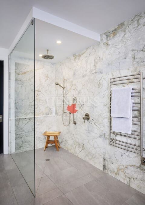 Spacious walk-in shower with marble walls, rain shower head, handheld sprayer, and towel warmer. Includes accessibility features and a small wooden bench