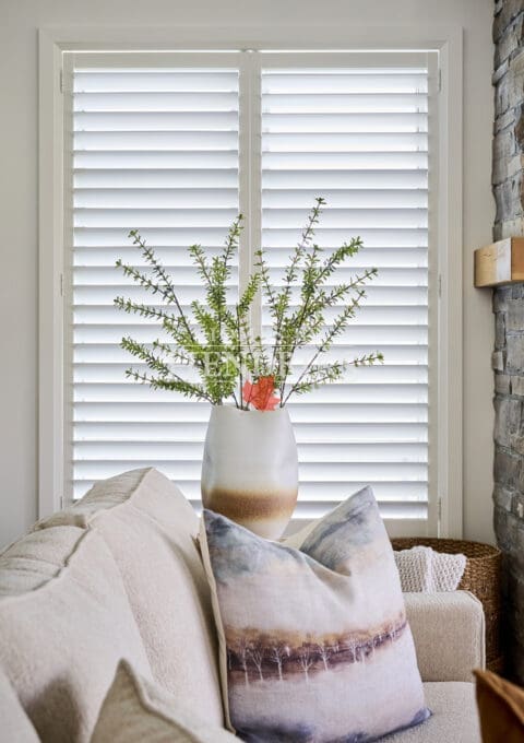 Soft neutral sofa with landscape-print pillows and a gradient vase of greenery set against white plantation shutters.