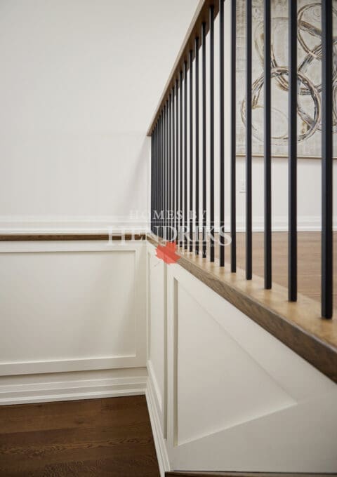 Elegant stairwell featuring white paneled walls, warm oak railing, and slim black spindles — a hallmark of refined transitional design by Homes By Hendriks.