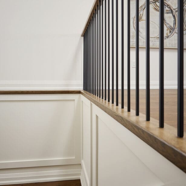 Elegant stairwell featuring white paneled walls, warm oak railing, and slim black spindles — a hallmark of refined transitional design by Homes By Hendriks.