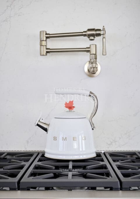 White SMEG kettle atop a stainless gas range with wall-mounted pot filler and marble backsplash for a balanced mix of function and luxury.
