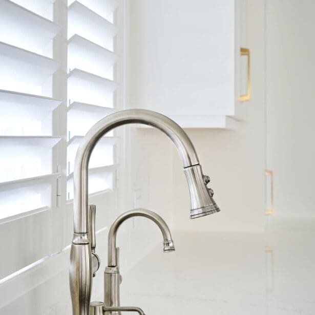 Brushed nickel kitchen faucet and matching prep tap in front of plantation shutters, blending modern function with timeless finishes.