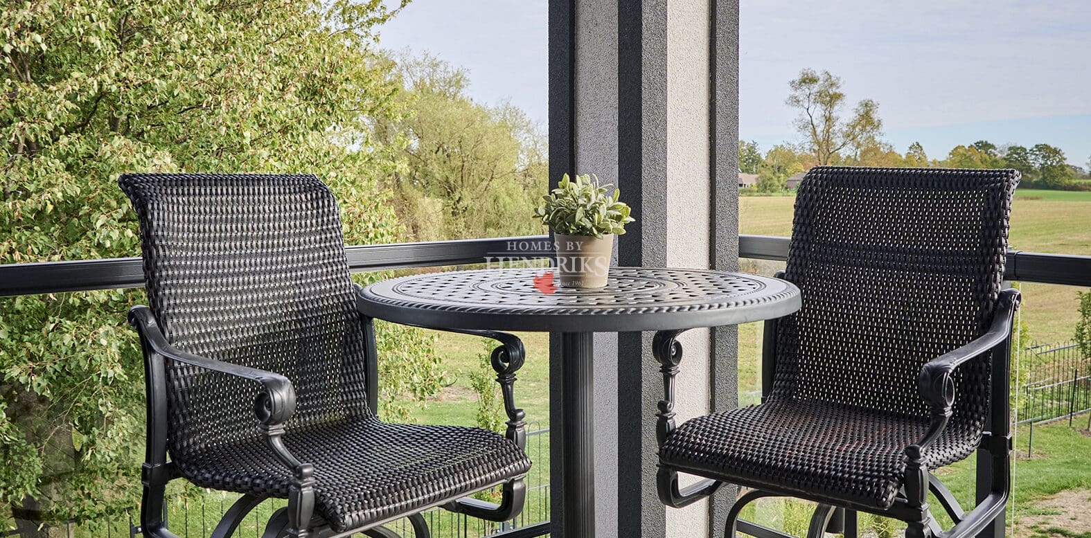 Modern balcony with black woven swivel bar chairs and a round metal bistro table, overlooking lush green trees and open fields.