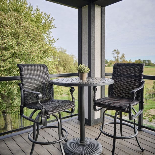 Modern balcony with black woven swivel bar chairs and a round metal bistro table, overlooking lush green trees and open fields.
