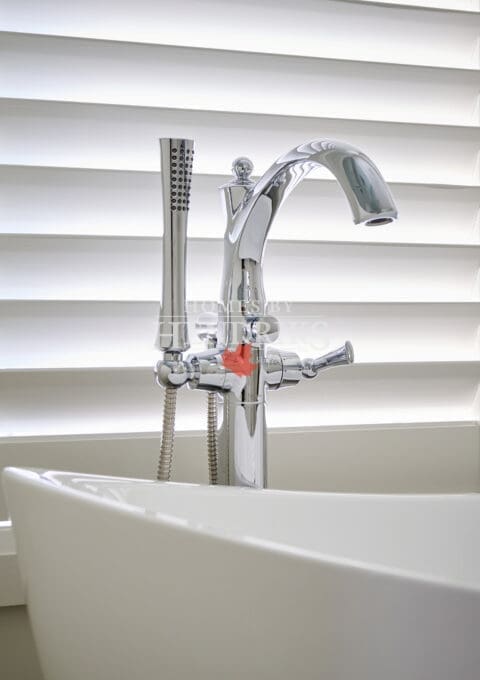 Polished chrome freestanding tub filler with handheld shower attachment set against soft white blinds, emphasizing clean modern luxury.