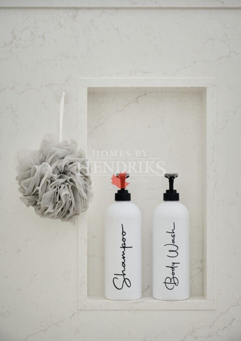 Marble shower niche with labeled white bottles and a neutral bath pouf, styled for a clean and timeless spa aesthetic