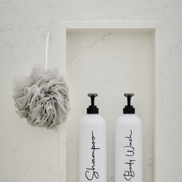 Marble shower niche with labeled white bottles and a neutral bath pouf, styled for a clean and timeless spa aesthetic