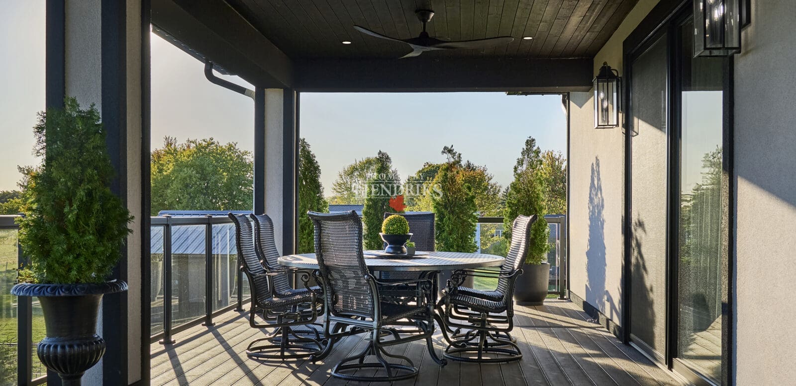 Spacious covered terrace with black wicker dining chairs, round table, and wood ceiling featuring recessed lighting and a ceiling fan, overlooking landscaped greenery.