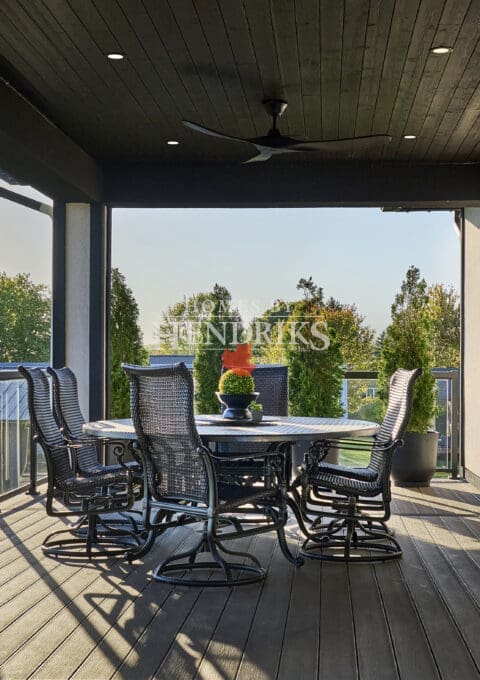Spacious covered terrace with black wicker dining chairs, round table, and wood ceiling featuring recessed lighting and a ceiling fan, overlooking landscaped greenery.