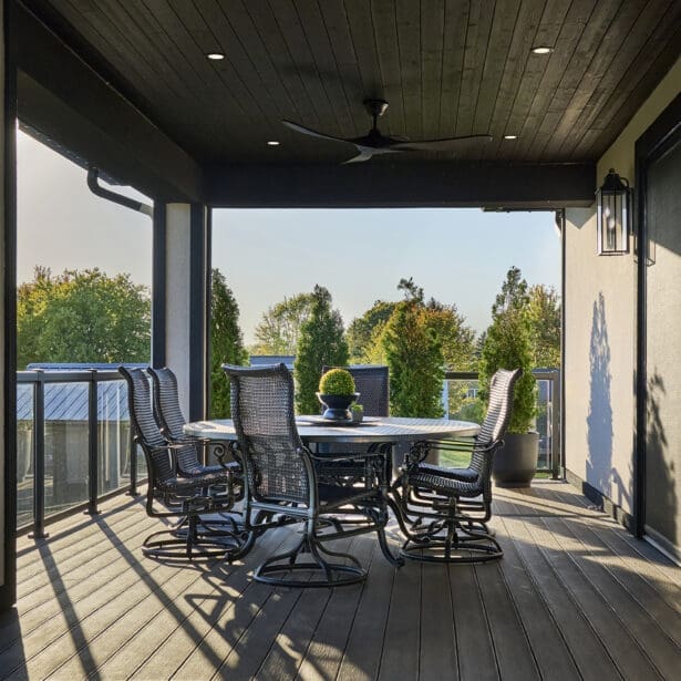 Spacious covered terrace with black wicker dining chairs, round table, and wood ceiling featuring recessed lighting and a ceiling fan, overlooking landscaped greenery.