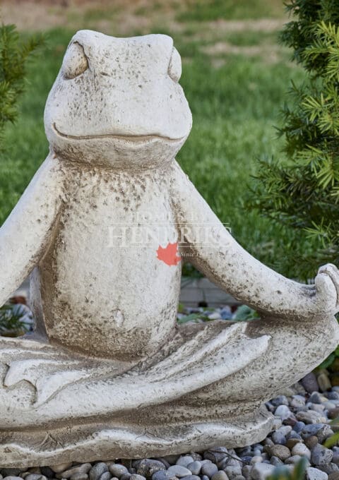 Whimsical stone frog sculpture seated in a meditative pose among garden greenery and decorative river stones.