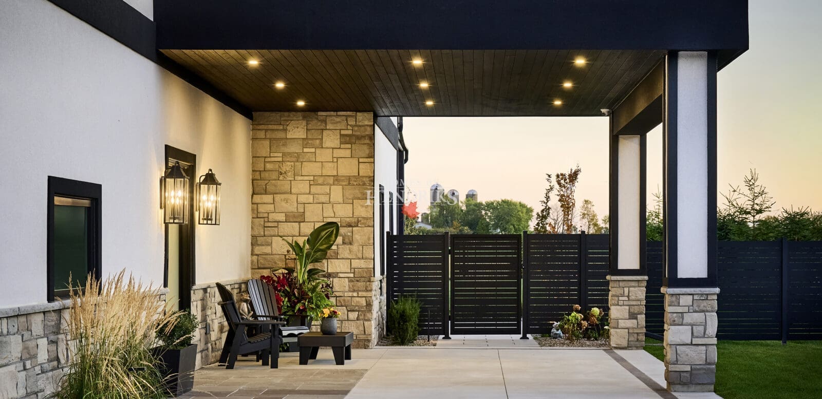 Covered entrance with wood soffit, recessed lighting, stone cladding, and modern black chairs surrounded by lush plantings and privacy fencing.