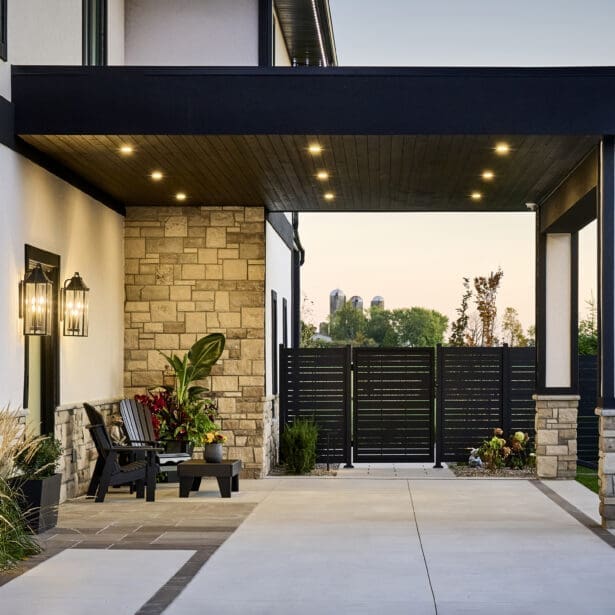 Covered entrance with wood soffit, recessed lighting, stone cladding, and modern black chairs surrounded by lush plantings and privacy fencing.