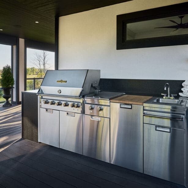 Covered terrace with stainless steel built-in grill, sink, and prep counter accented by stone-textured finishes and open countryside views.