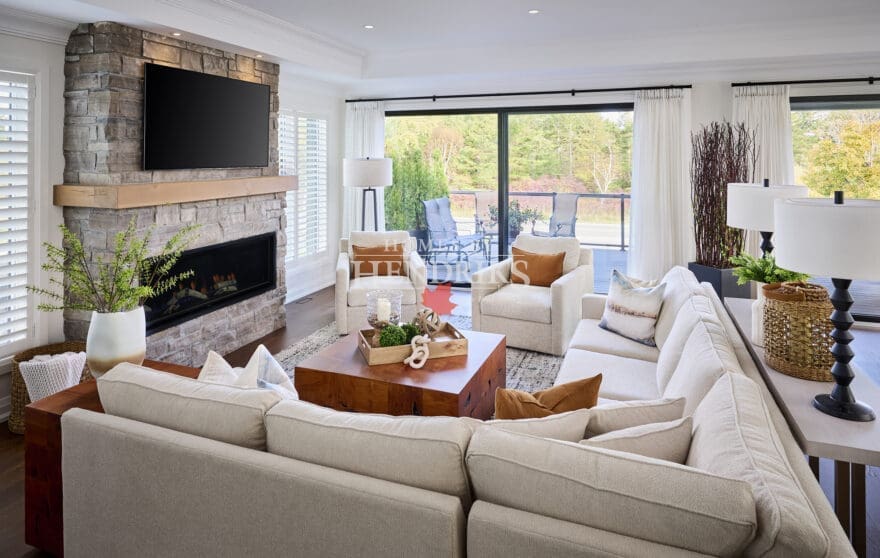 Bright open-concept living room featuring a stone fireplace with wood mantel, sectional seating, and soft neutral décor overlooking an outdoor patio.