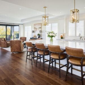 Spacious kitchen and living area with wide-plank hardwood floors, white cabinetry, gold pendant lighting, and caramel leather counter stools.