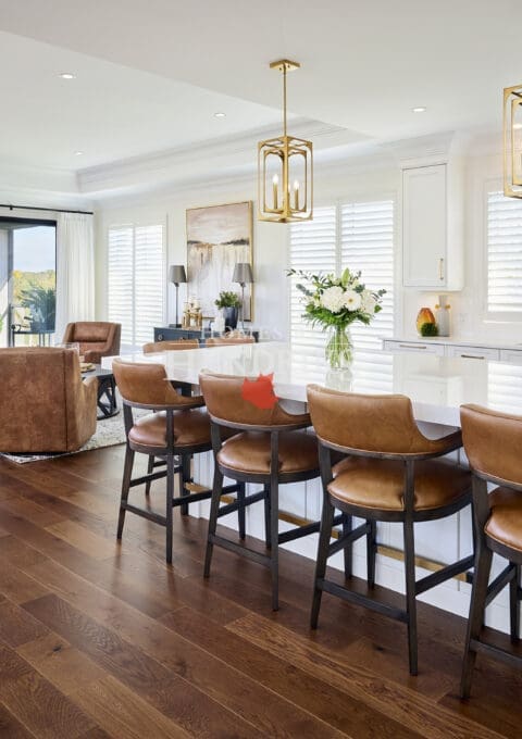 Spacious kitchen and living area with wide-plank hardwood floors, white cabinetry, gold pendant lighting, and caramel leather counter stools.