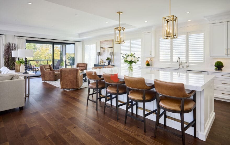 Spacious kitchen and living area with wide-plank hardwood floors, white cabinetry, gold pendant lighting, and caramel leather counter stools.