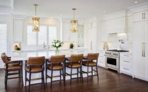 Bright modern kitchen featuring white shaker cabinetry, gold hardware, a large quartz island, and leather counter stools with dark wood frames.