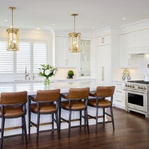 Bright modern kitchen featuring white shaker cabinetry, gold hardware, a large quartz island, and leather counter stools with dark wood frames.