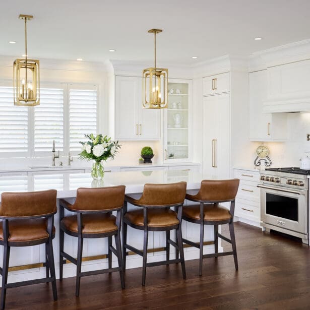 Bright modern kitchen featuring white shaker cabinetry, gold hardware, a large quartz island, and leather counter stools with dark wood frames.