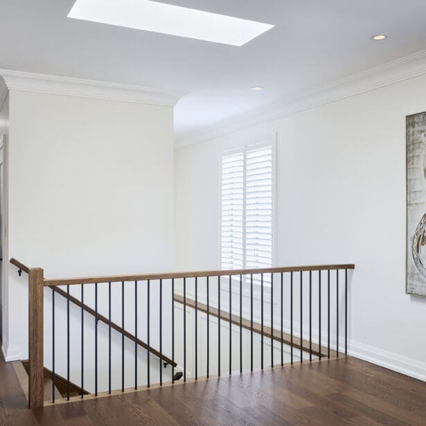 Open upper hallway with oak flooring, white walls, black railing, and oversized abstract artwork beneath a skylight.